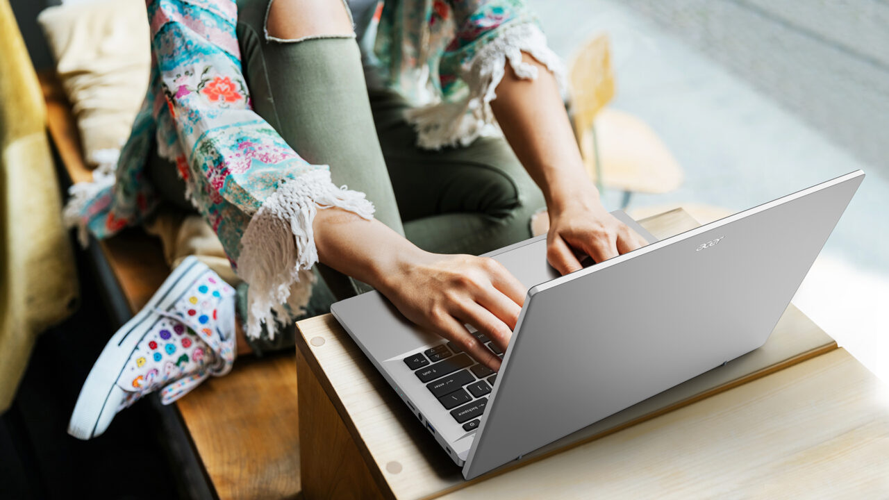 Kobieta w kolorowych ubraniach siedzi przy drewnianym stole i pracuje na laptopie.