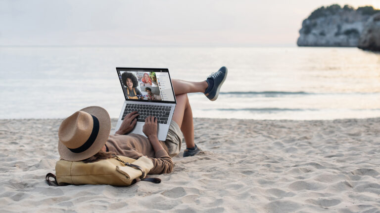 Osoba leżąca na brzuchu na plaży z laptopem, na którym widać rozmowę wideo z trzema osobami, w tle morze i skały.