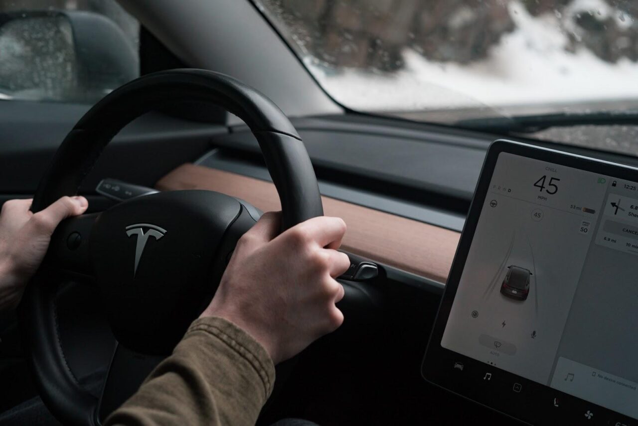 Zasięg Tesli. Wnętrze samochodu Tesla z widokiem na kierownicę i ręce kierowcy oraz duży ekran dotykowy z wyświetlonym interfejsem pojazdu.