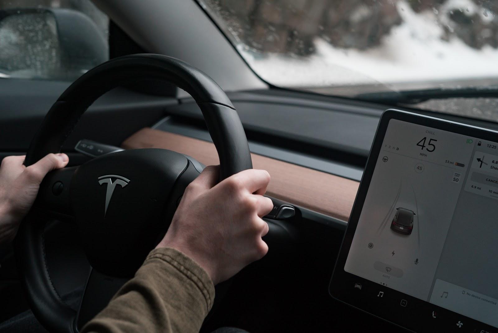 Wnętrze samochodu Tesla z widokiem na kierownicę i ręce kierowcy oraz duży ekran dotykowy z wyświetlonym interfejsem pojazdu.