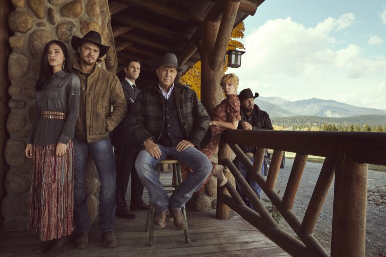 Grupa sześciu osób w stylu westernowym, stojąca na drewnianym werandzie górskiej chaty, z górskim krajobrazem w tle.