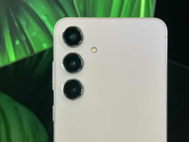 Tylna część smartfona w kolorze białym z potrójnym systemem kamer i diodą LED, na tle zielonych liści.