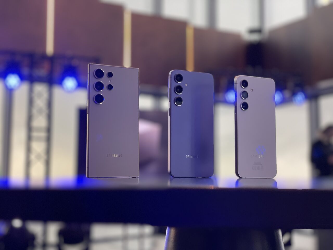 Trzy smartfony Samsung ustawione jeden za drugim na rozmytym tle z niebieskimi światłami.