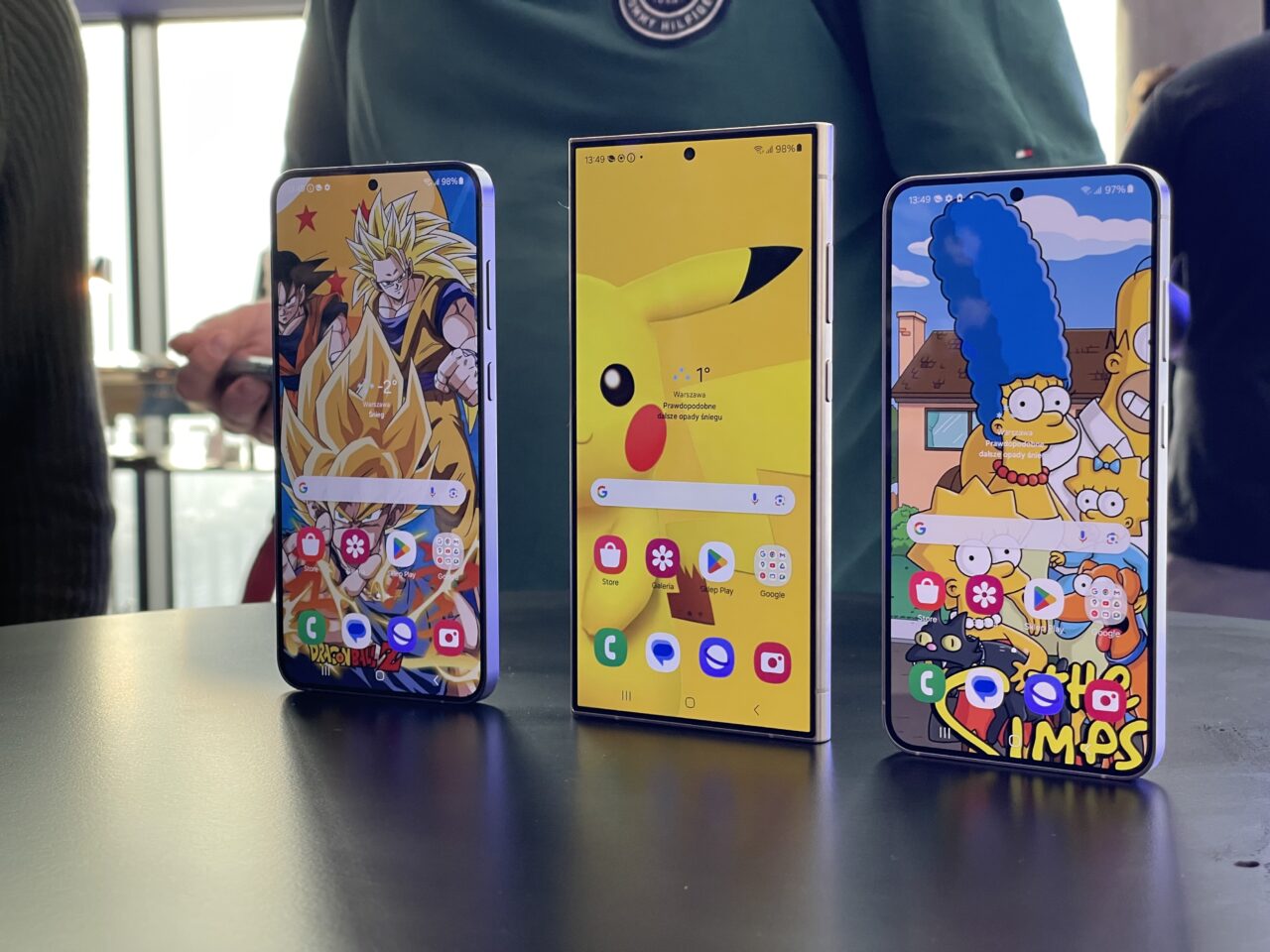 Trzy smartfony ustawione obok siebie wyświetlają różne kolorowe tapety z postaciami z animacji: Dragon Ball, Pokémon i The Simpsons.