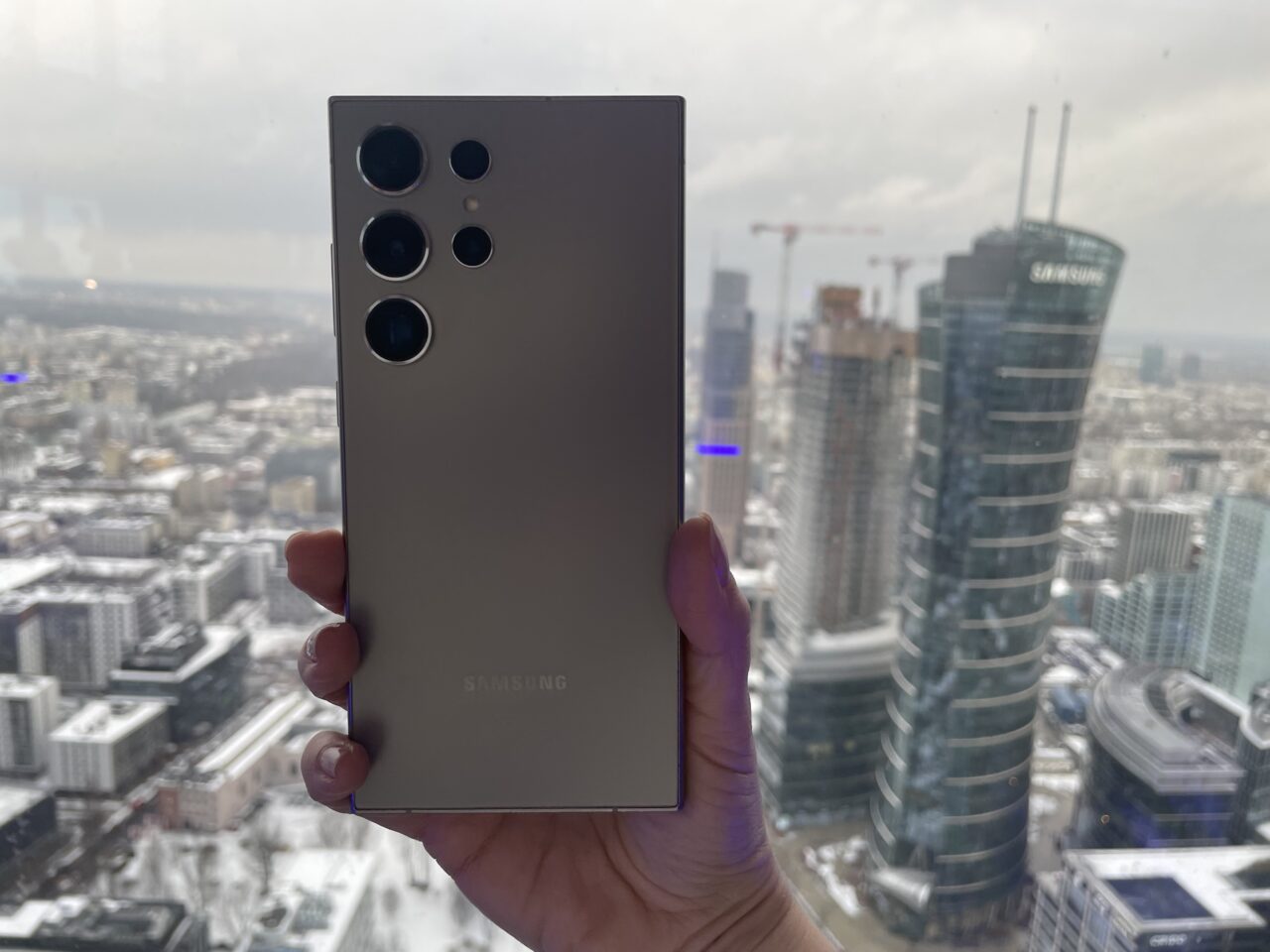 Tylna kamera smartfona marki Samsung w kolorze srebrnym trzymana na tle panoramicznego widoku na miejski krajobraz z wysokich budynków.
