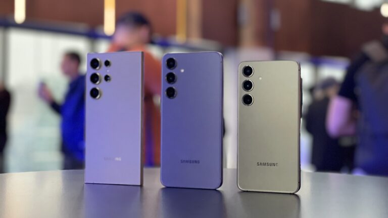 Trzy smartfony Samsung ustawione pionowo na stole z rozmytym tłem, gdzie widać sylwetki ludzi.