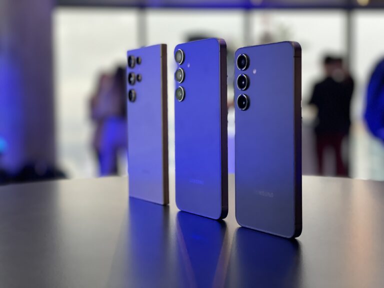 Trzy smartfony Samsung ustawione obok siebie na błyszczącym blacie, z tylnymi aparatami skierowanymi w stronę obserwatora, na rozmytym tle z ludźmi.