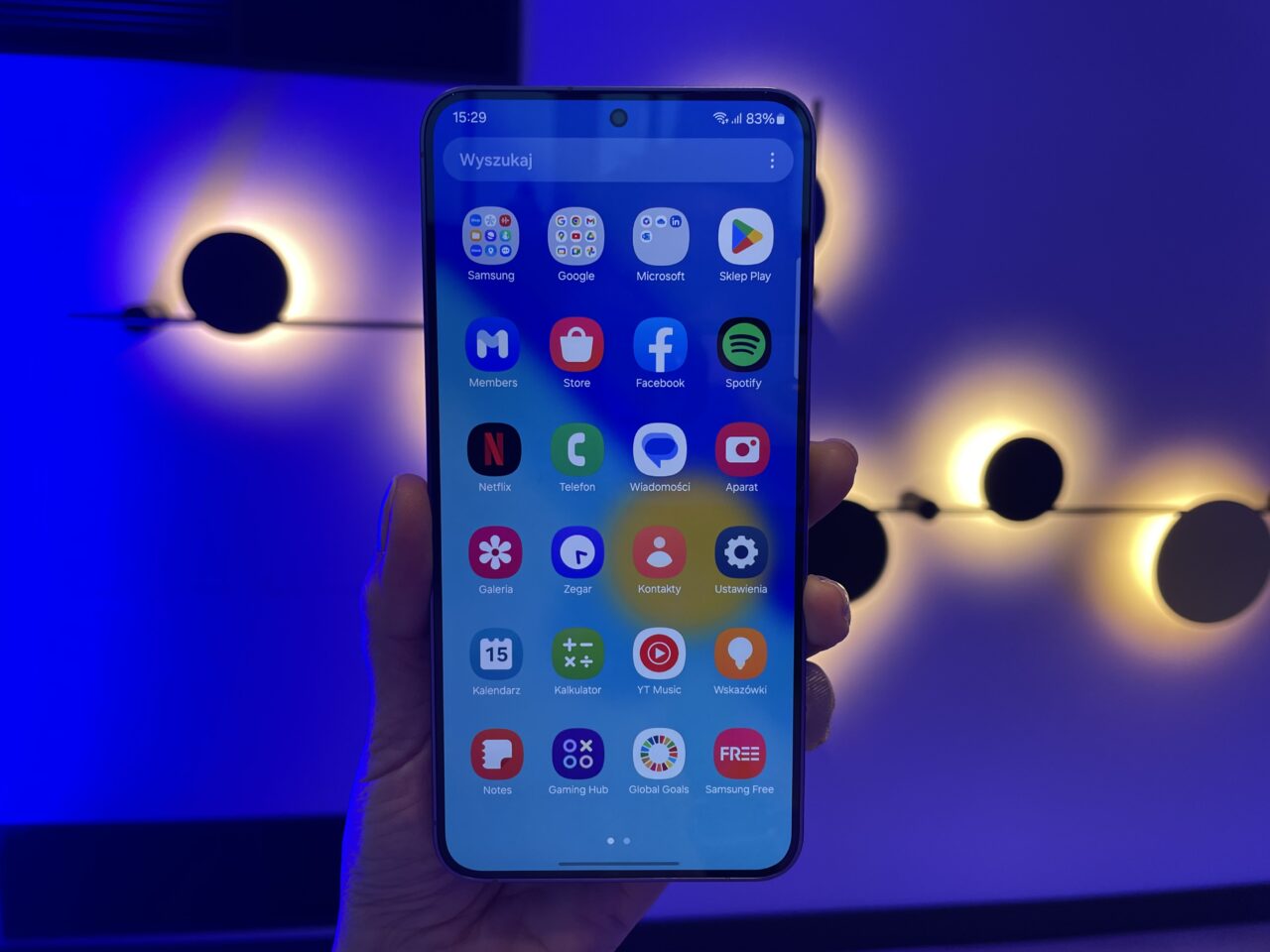 Ekran smartfona Samsung Galaxy S24 Plus wyświetlający kolorowe ikony aplikacji trzymany w ręce na tle niebieskiej ściany z rozproszonymi żółtymi światłami.