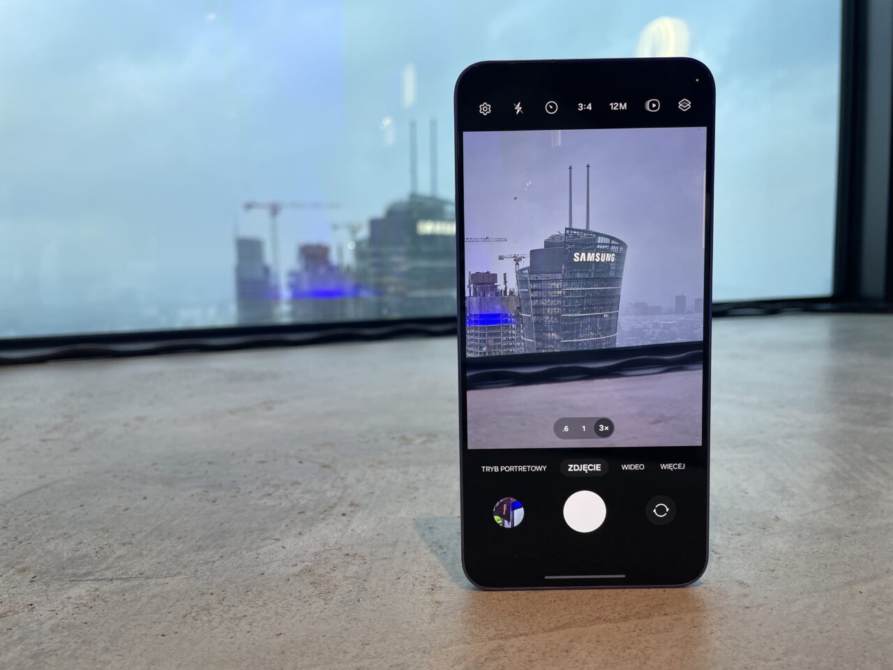 Smartfon leżący na blacie, z ekranem pokazującym widok na budynek z logo firmy Samsung, uchwycony przez ekran smartfona.
