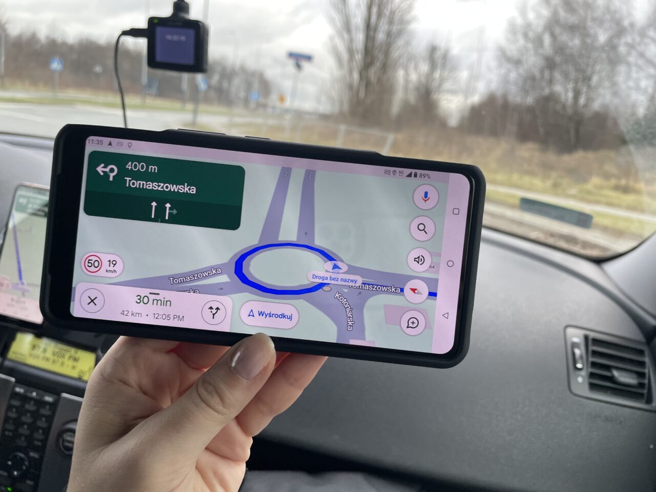 Ręka trzymająca smartfon z włączoną aplikacją nawigacyjną w samochodzie, pokazującą mapę i wskazówki dojazdu, z nazwą ulicy Tomaszowska i czasem przyjazdu 30 minut.