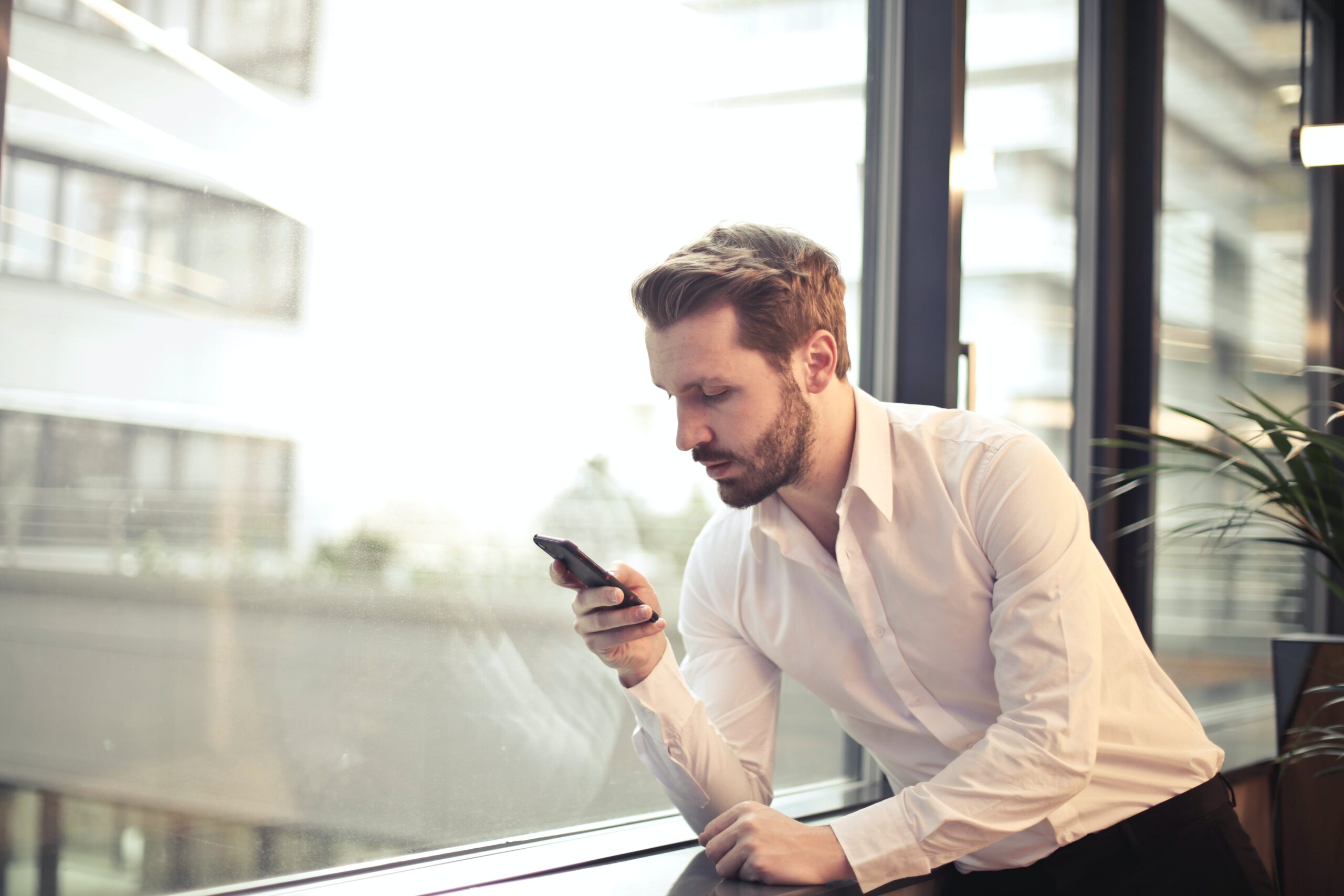 Mężczyzna w białej koszuli siedzący obok okna i patrzący na telefon komórkowy.