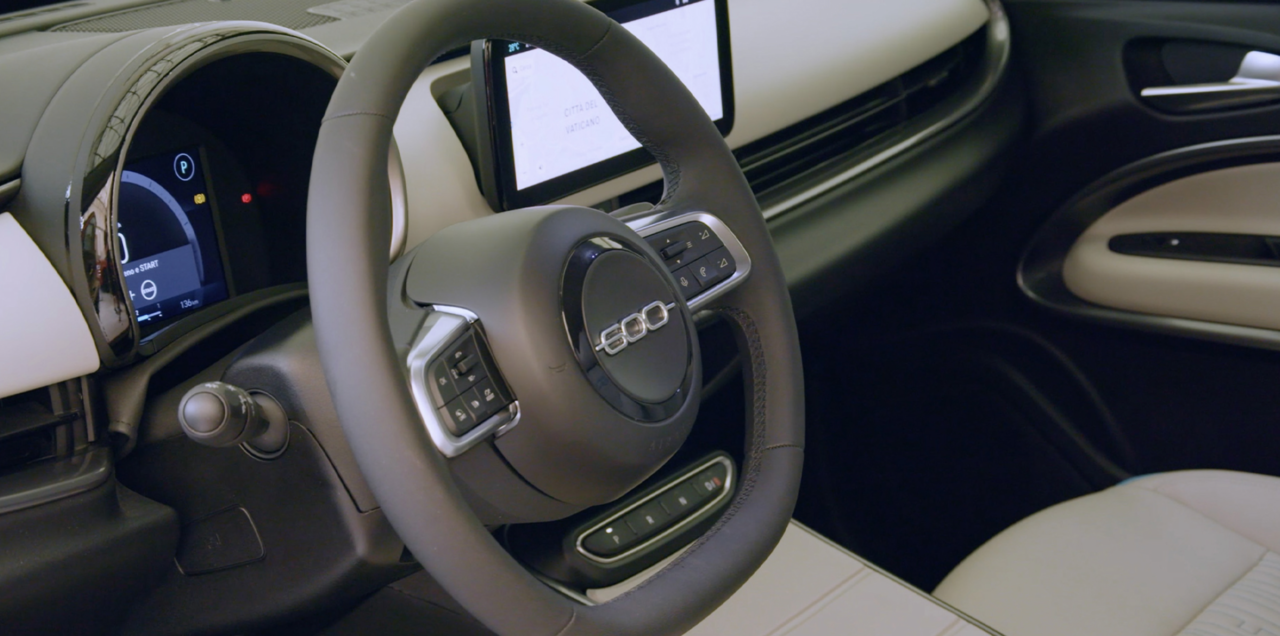Wnętrze samochodu z widokiem na kierownicę z logiem 600, cyfrowymi zegarami i częścią deski rozdzielczej z ekranem nawigacji.