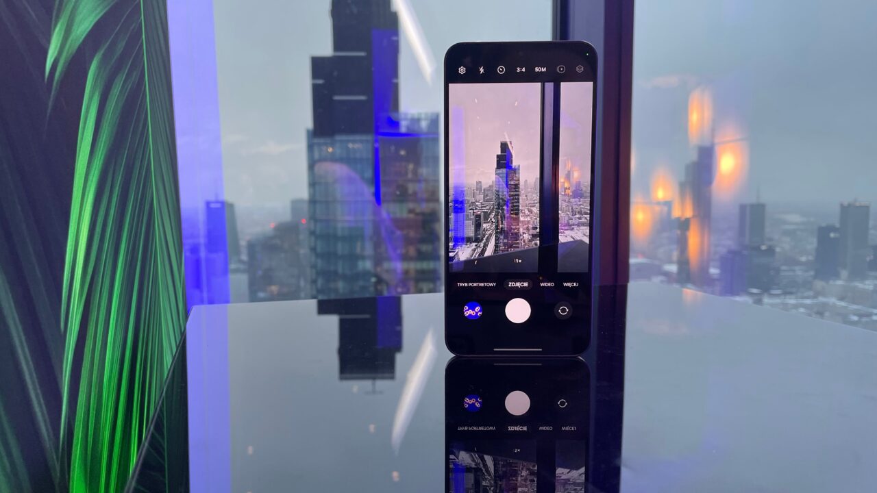 Smartfon trzymany przed panoramą miejską, robiący zdjęcie drapaczy chmur przez szklaną szybę z odbiciem świateł wewnątrz pomieszczenia.