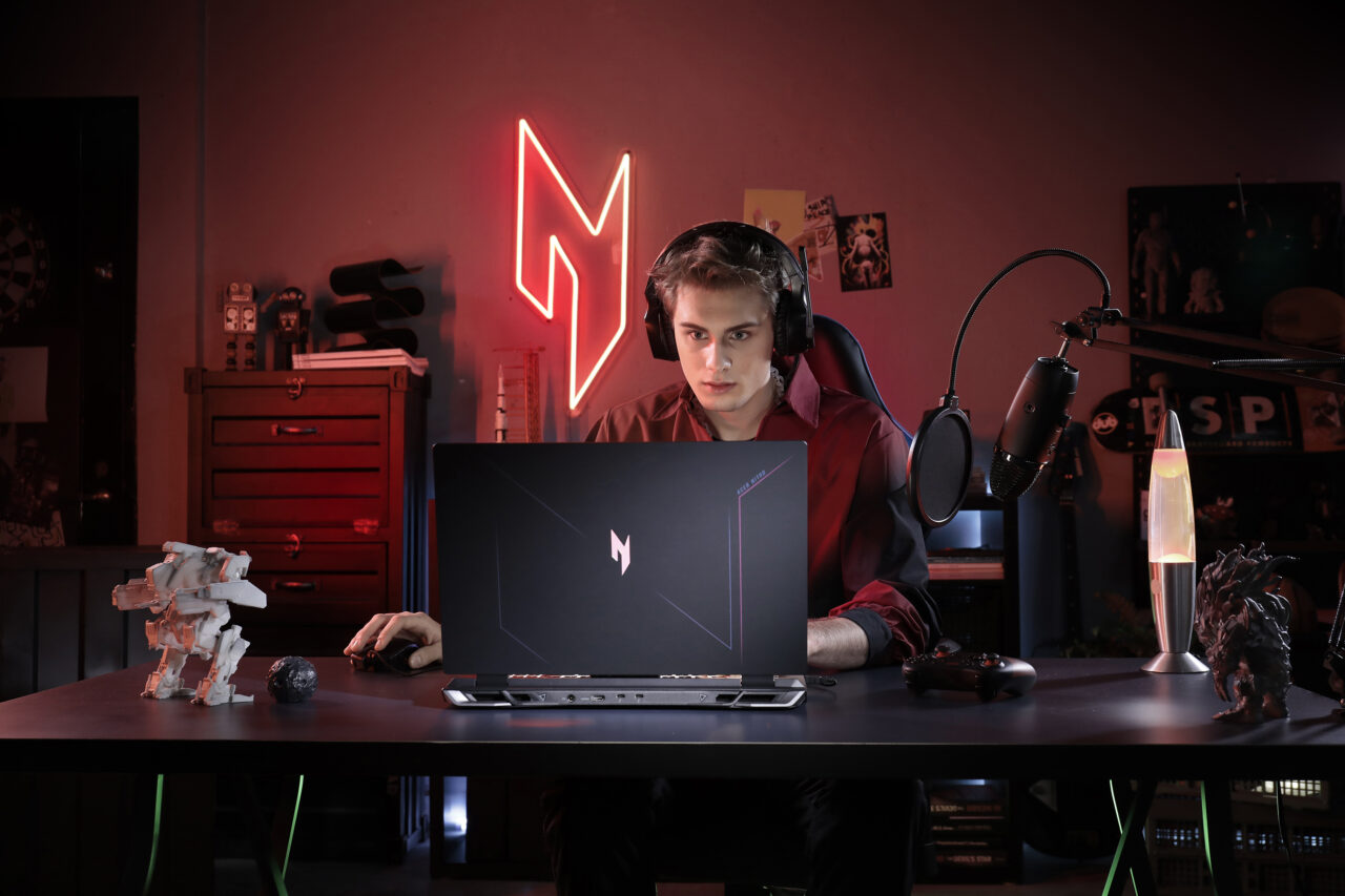 Osoba grająca w gry na komputerze z otwartym laptopem na biurku, nosząca słuchawki, w otoczeniu dekoracji związanych z grami i czerwonym neonem w kształcie litery M na ścianie.