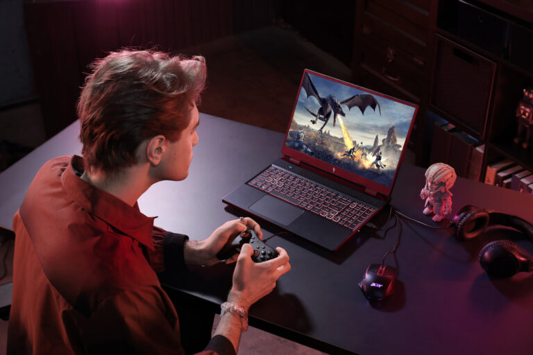 Młody mężczyzna grający w grę wideo na laptopie z grafiką przedstawiającą smoka i postacie fantastyczne, na stole obok figurka i słuchawki gamingowe.