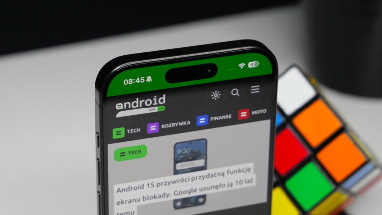 Apple iPhone 15 Pro z otwartą stroną internetową o tematyce Android, położony obok kostki Rubika, na tle z rozmytymi elementami.