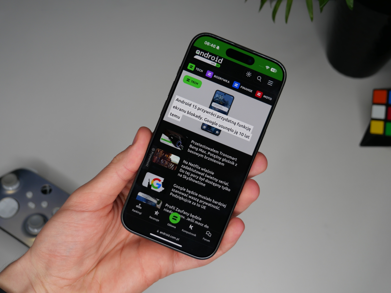 Osoba trzymająca iPhone 15 Pro wyświetlający stronę internetową z artykułami o tematyce Android, z widocznym logo serwisu i zielonym paskiem stanu u góry ekranu.