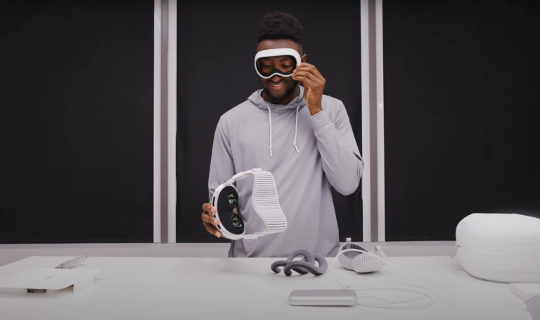 Mężczyzna w ciemnym pomieszczeniu prezentuje futurystyczne gogle VR Apple Vision Pro, trzymając je w prawej ręce; nosi on także pasującą do gogli, sportową opaskę na oczy i stylową, szarą bluzę z kapturem. Na białym stole obok znajdują się dodatkowe akcesoria związane z goglami VR.