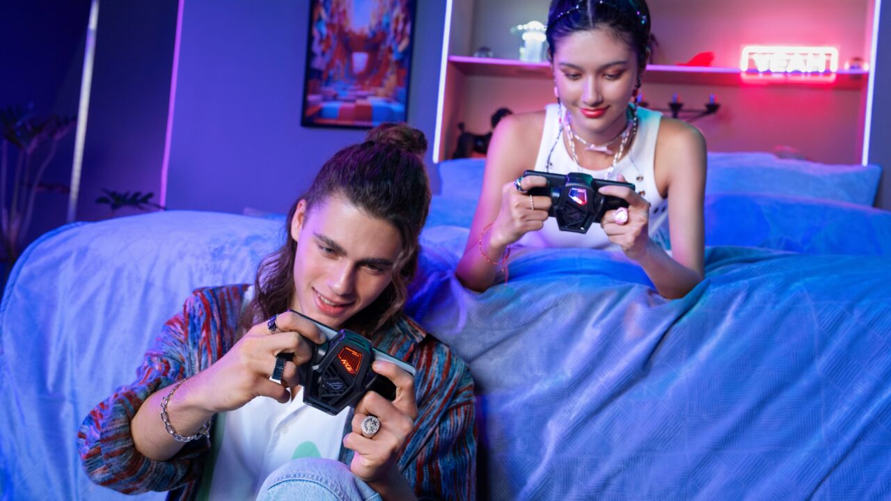 Dwoje młodych ludzi gra w gry wideo na kanapie w pokoju z neonowym oświetleniem. W rękach mają smartfony Asus ROG Phone 8 Pro i Activecooler