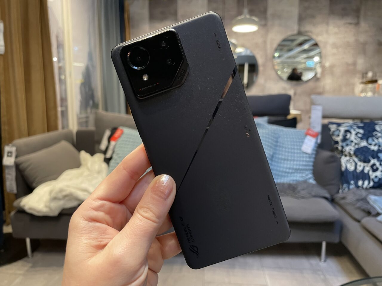 Czarny smartfon trzymany w dłoni z tyłu aparatu na tle wnętrza salonu.