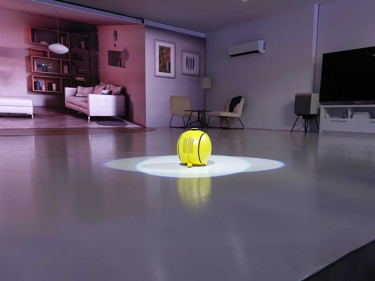 Nowoczesny salon z żółtym robotem Samsung Ballie na środku jaśniejszej okrągłej powierzchni na podłodze, meble w odcieniach szarości i beżu, klimatyzacja i telewizor na ścianie.