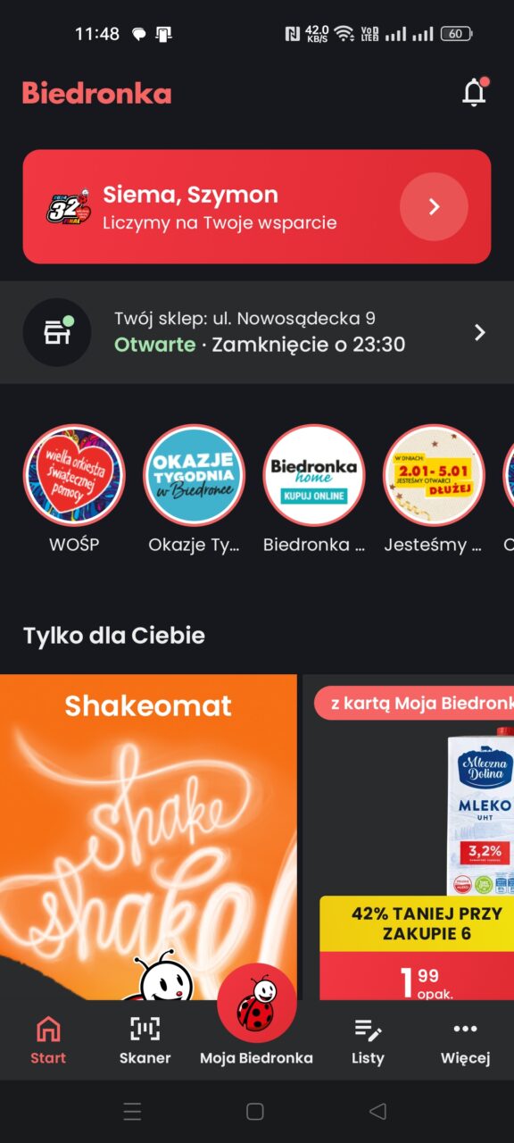 Zrzut ekranu z aplikacji Biedronka na smartfonie, pokazujący powitanie użytkownika o imieniu Szymon, opcje zakupów online, oraz różne promocje, w tym Shakeomat i obniżoną cenę mleka przy zakupie sześciu opakowań. Na dole ekranu widoczne są ikony nawigacji aplikacji.