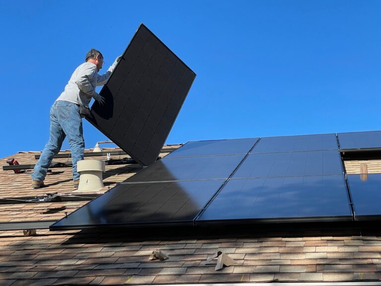 Mężczyzna instalujący panele słoneczne na dachu domu pod błękitnym niebem.