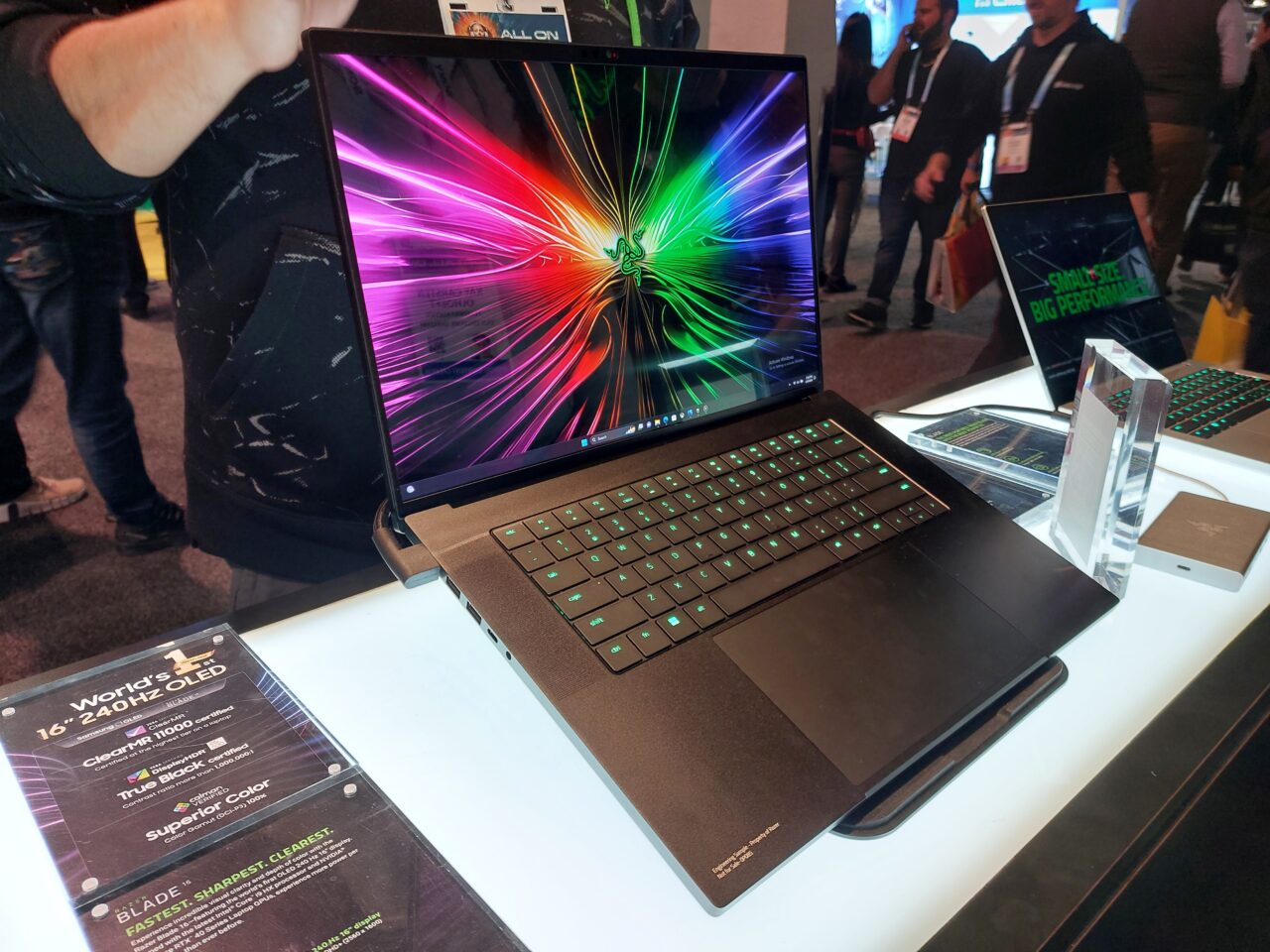 Laptop Razer Blade 16 z ekranem OLED o przekątnej 16 cali i rozdzielczości 4K prezentowany na wystawie, z otwartą klapą i widocznym kolorowym obrazem na ekranie. Na pierwszym planie tabliczka informacyjna o cechach ekranu, w tle inne urządzenia i odwiedzający.