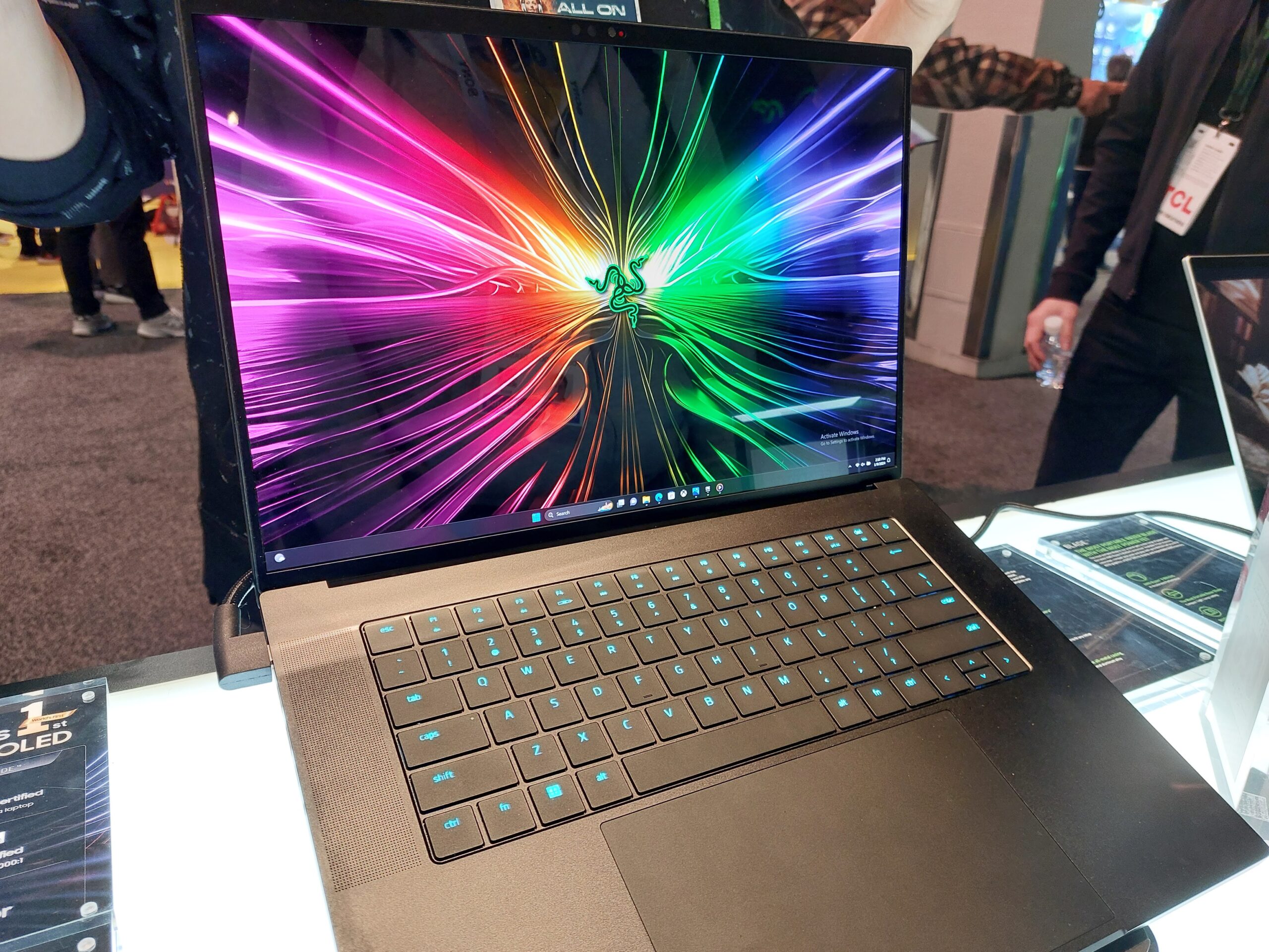 Laptop Razer Blade 16 z kolorową tapetą z efektem świetlnym na wyświetlaczu, wystawiony na stoisku, w tle osoby rozmyte w ruchu.