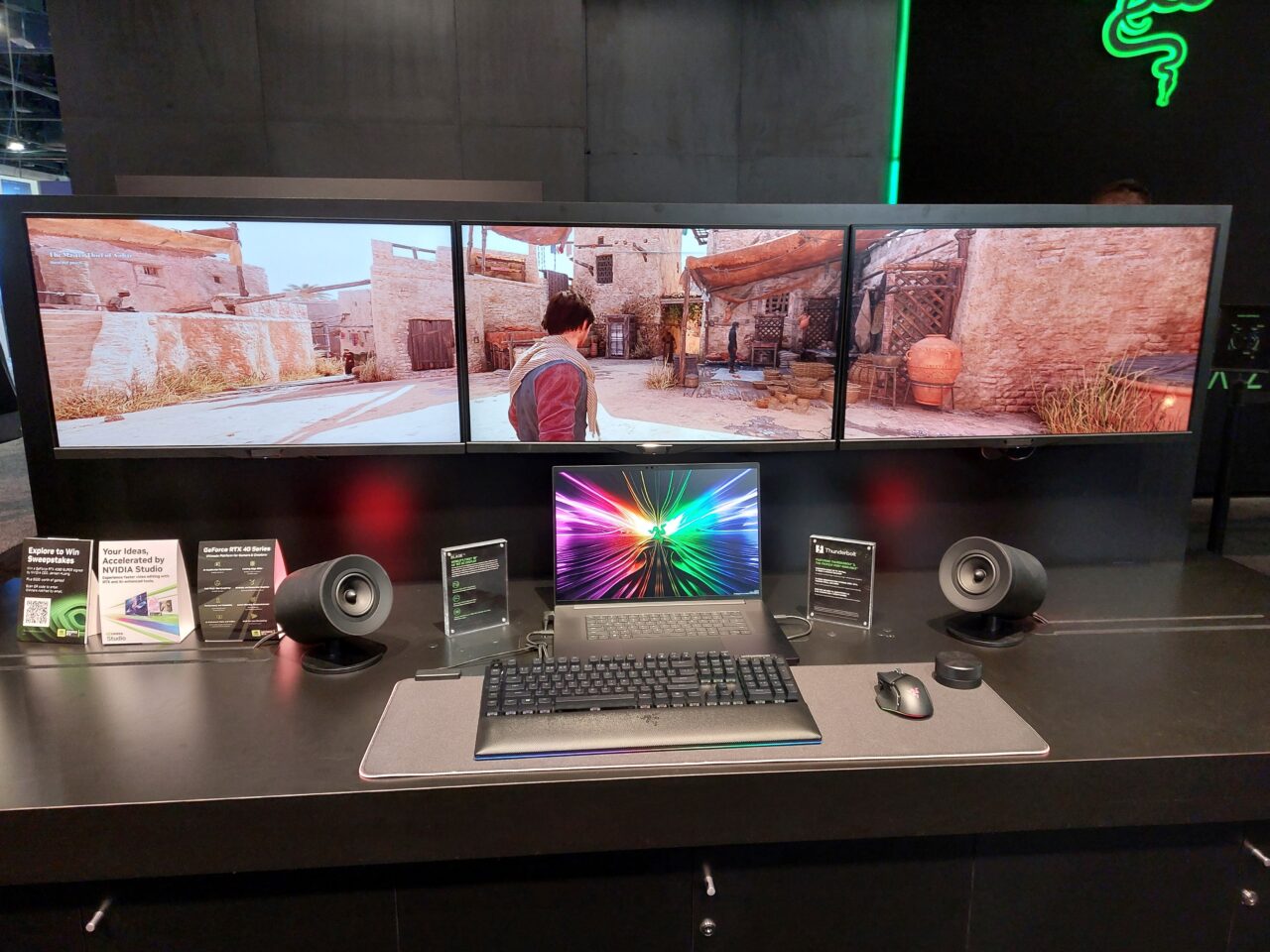 Zestaw komputerowy z laptopem Razor Blade 18 i  trzema monitorami wyświetlającymi grę oraz  myszką, głośnikami na wystawowym stole.