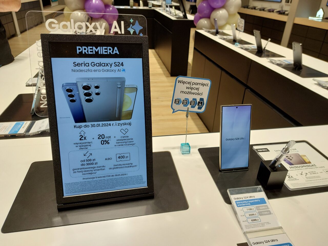 Wnętrze sklepu z elektroniką z reklamą telefonu Samsung Galaxy S24 i wystawionymi modelami telefonów na tle balonów.