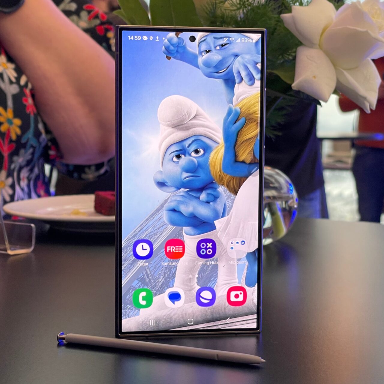 Smartfon stoi pionowo na stole z wyświetlanym tapetem przedstawiającym dwie postacie z animacji, w tle nieostre elementy wnętrza.