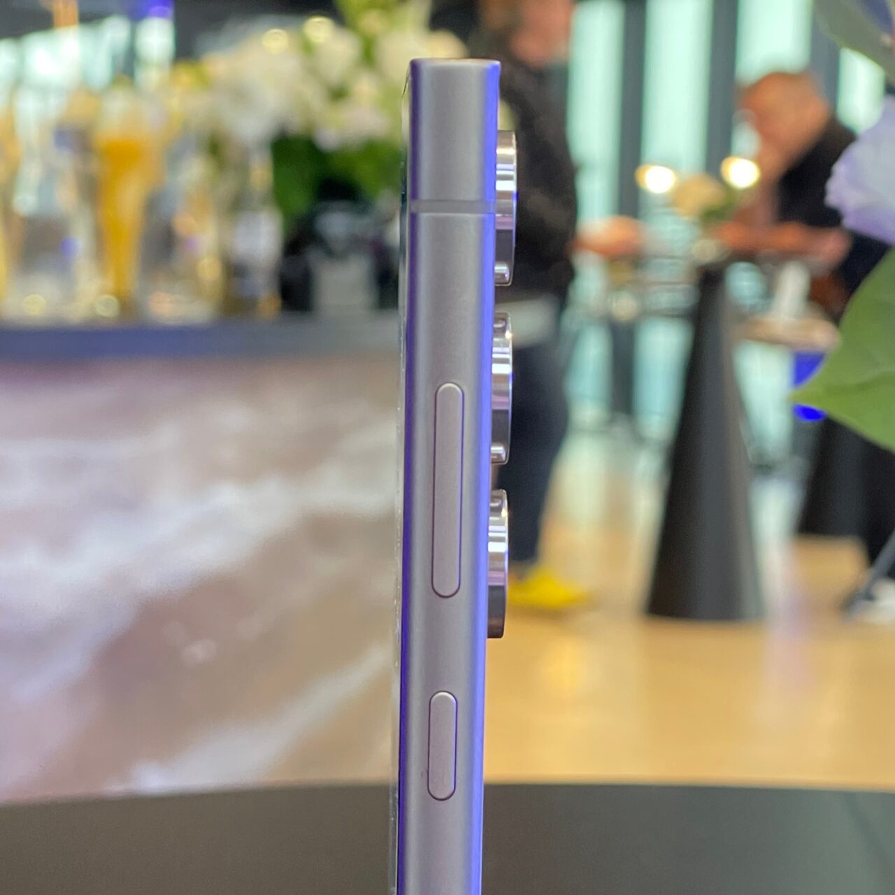Zbliżenie na fioletowy smartphone stojący pionowo na stole z rozmytym tłem, na którym widać ludzi i bar.