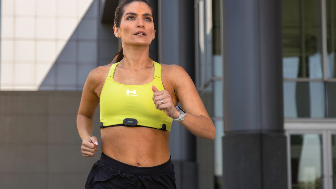Kobieta biega na zewnątrz w żółtym topie sportowym, czarnych szortach, z pulsometrem na klatce piersiowej i zegarkiem sportowym na nadgarstku.