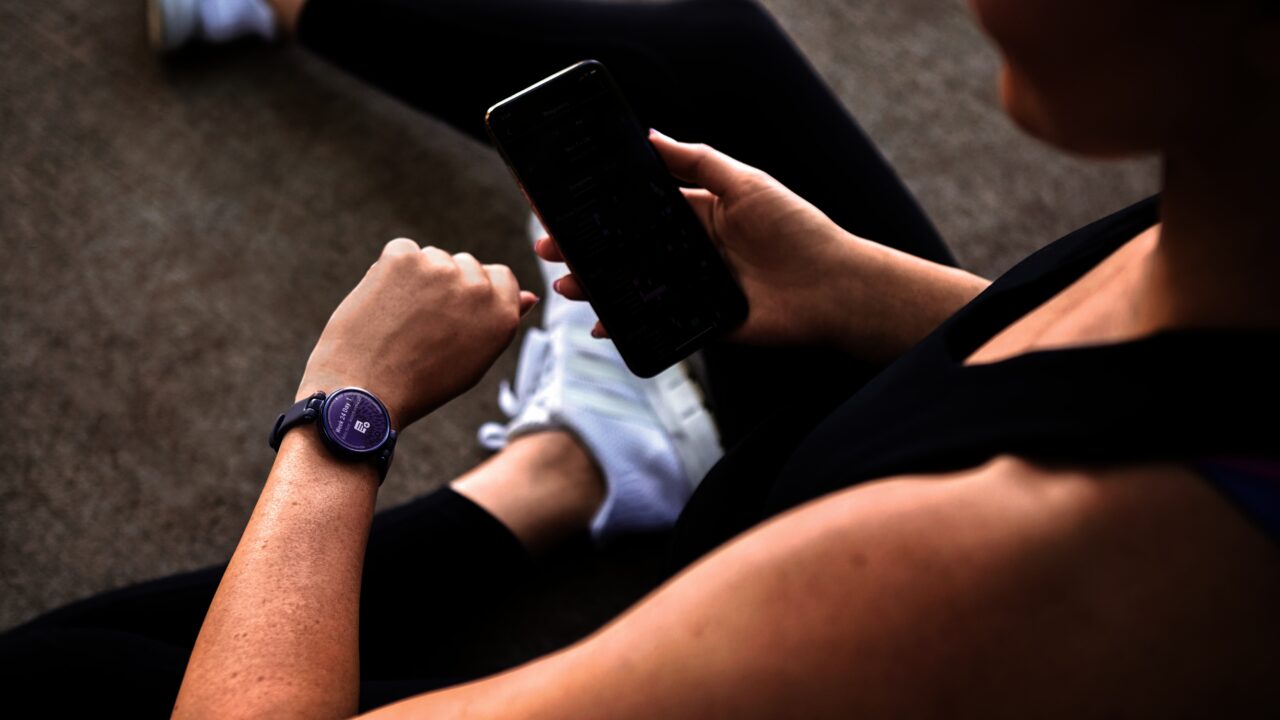 Osoba siedząca na podłodze sprawdza aplikację na smartfonie trzymając go w prawej dłoni, na lewym nadgarstku ma inteligentny zegarek; widok z profilu, nacisk na ręce i urządzenia.