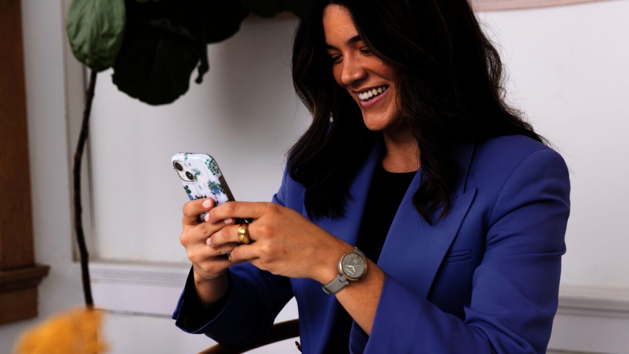 Uśmiechnięta kobieta w niebieskim garniturze trzymająca smartfon z kolorowym etui.