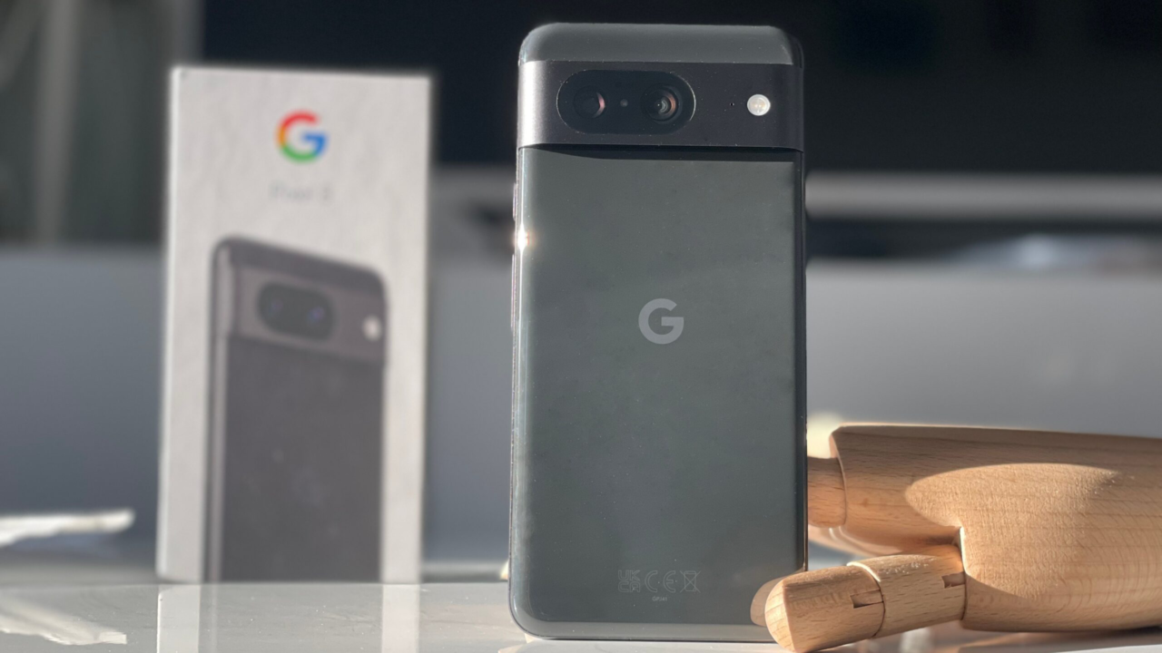 Smartfon Google Pixel z tyłu oparty o drewnianą figurę, z rozmytym pudełkiem z logiem Google w tle.