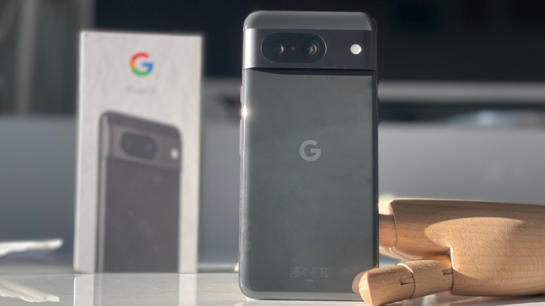 Smartfon Google Pixel 6 stojący pionowo na stole z rozmytym tłem i kartonowym pudełkiem tego telefonu w tle po lewej stronie oraz drewniana figurka w prawym dolnym rogu zdjęcia.
