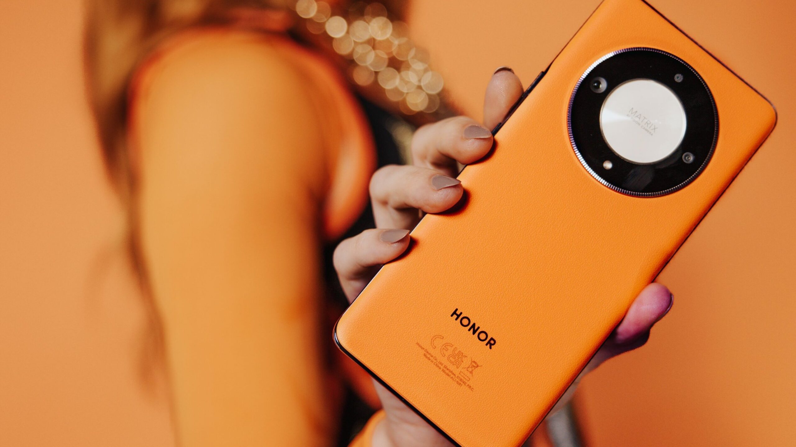 Osoba trzymająca pomarańczowy smartfon Honor z okrągłym modułem aparatu.