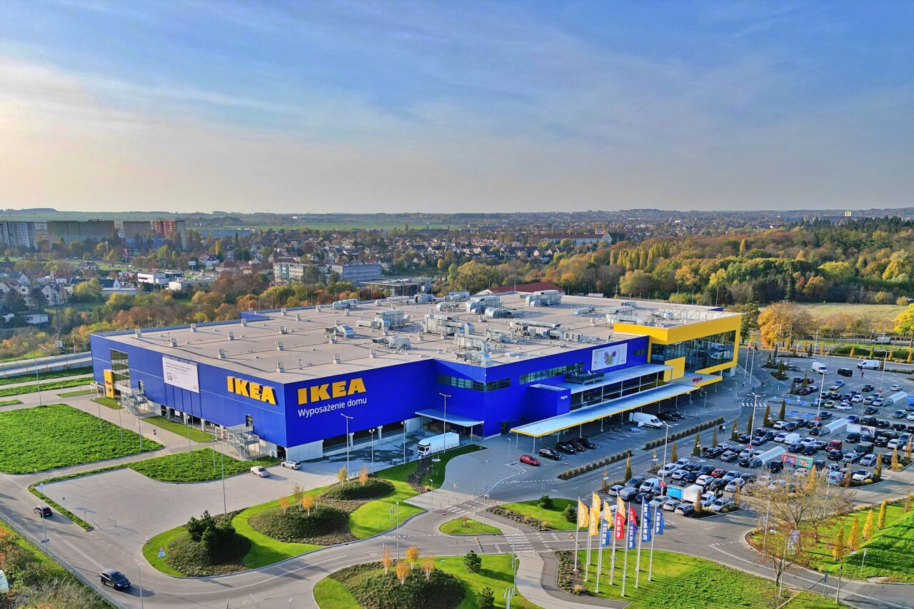 Widok z lotu ptaka na sklep IKEA z dużym parkingiem, flagami na masztach i zalesioną okolicą w tle.