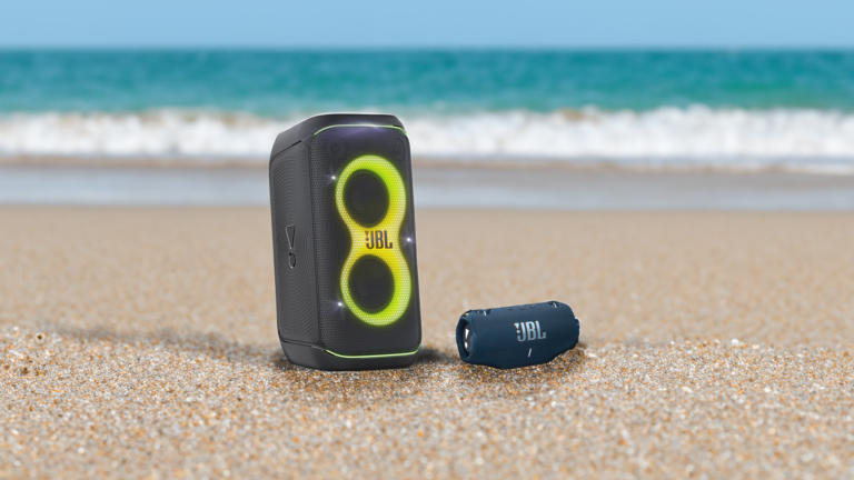Dwa głośniki przenośne marki JBL na piaszczystej plaży, większy głośnik stoi pionowo, a mniejszy leży poziomo, w tle morze i niebo.
