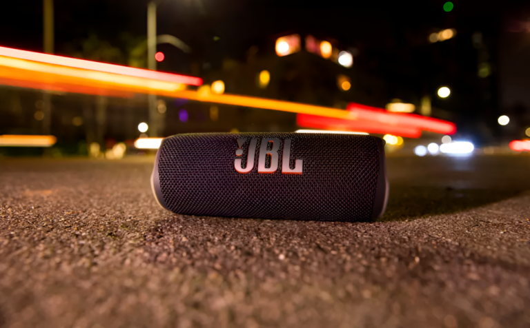 Przenośny głośnik JBL leżący na asfalcie nocą z rozmytymi światłami pojazdów w tle.
