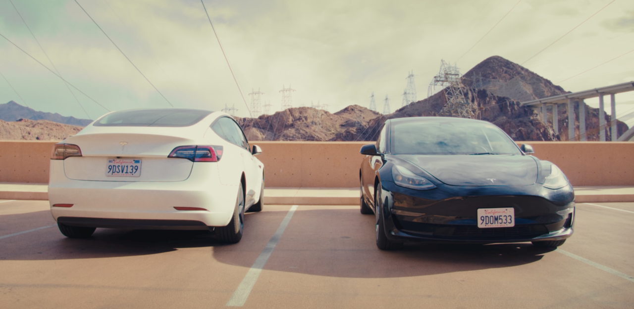 Białe i czarne auto marki Tesla zaparkowane na betonowym parkingu, w tle skaliste wzgórza i słupy wysokiego napięcia.