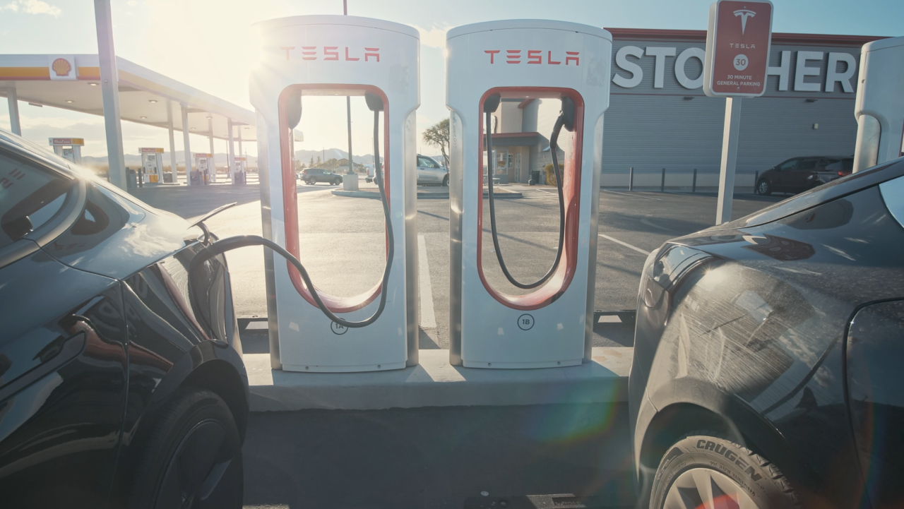 Stacja ładowania samochodów elektrycznych Tesla z zaparkowanym i podłączonym do ładowarki pojazdem, na tle słonecznego nieba.