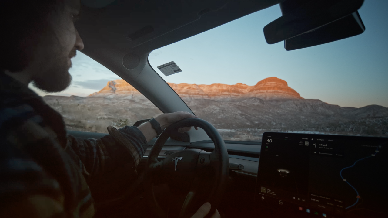 Mężczyzna prowadzi samochód elektryczny z widocznym logo marki na kierownicy, na tle zachodzącego słońca oświetlającego górski krajobraz, z centrum sterowania dotykowego na desce rozdzielczej.