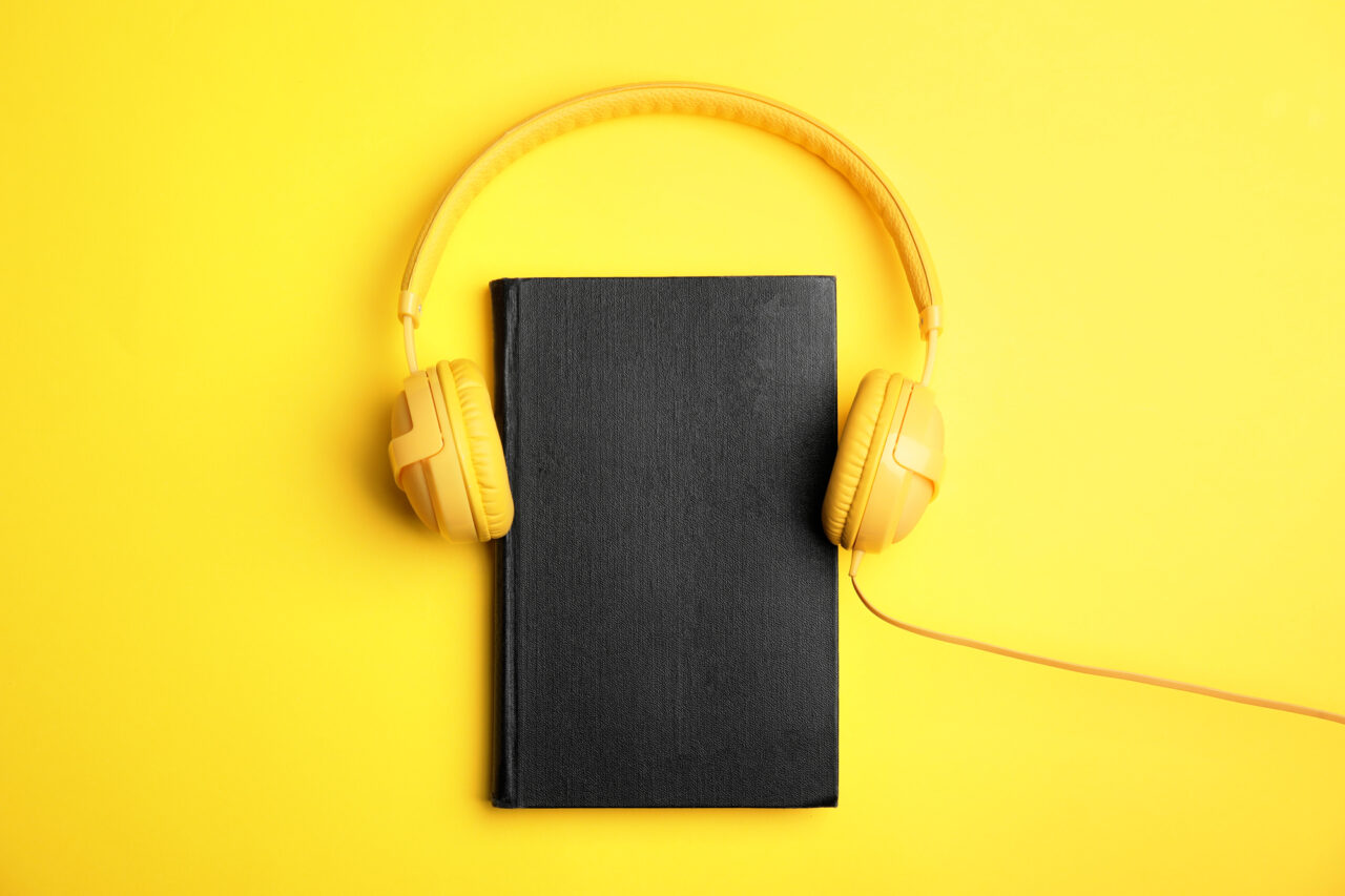 Zdjęcie ilustrujące audiobooki za darmo. Czarne, zamknięte książka leżąca na żółtym tle z nałożonymi żółtymi słuchawkami nauszynymi.