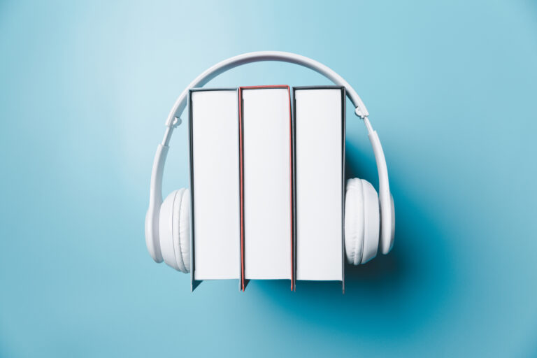 Trzy zamknięte książki ustawione pionowo z białymi słuchawkami nauszonymi na środkowej książce, na błękitnym tle.
