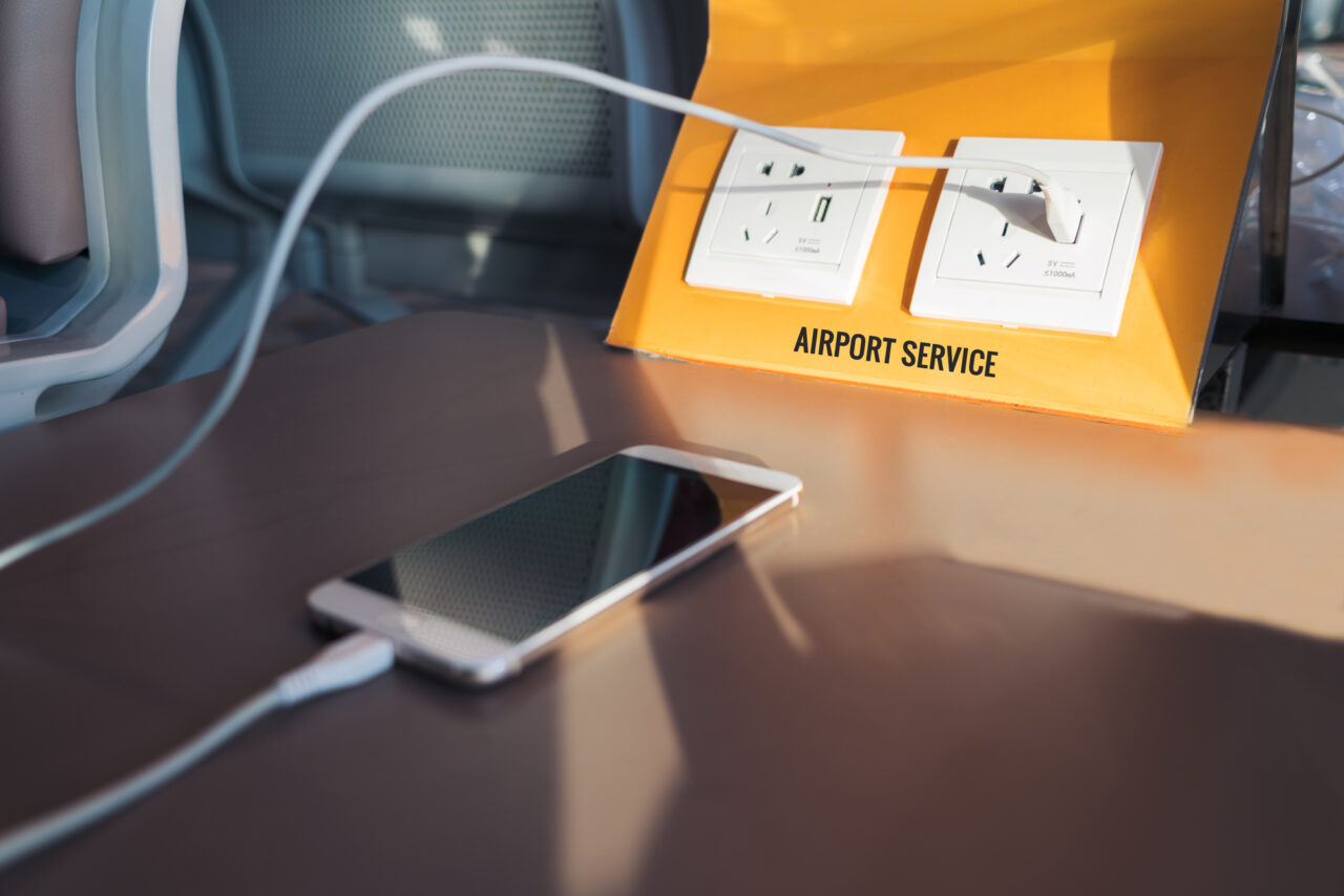 Zdjęcie do artykułu o juice jacking. Stacja ładowania na lotnisku z gniazdkami elektrycznymi i napisem "AIRPORT SERVICE", obok której znajduje się telefon komórkowy podłączony kablem.