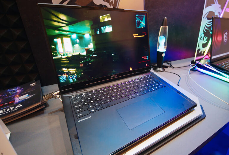 Laptop gamingowy MSI na biurku z otwartym oknem gry, obok lampa lawowa i drugi komputer.