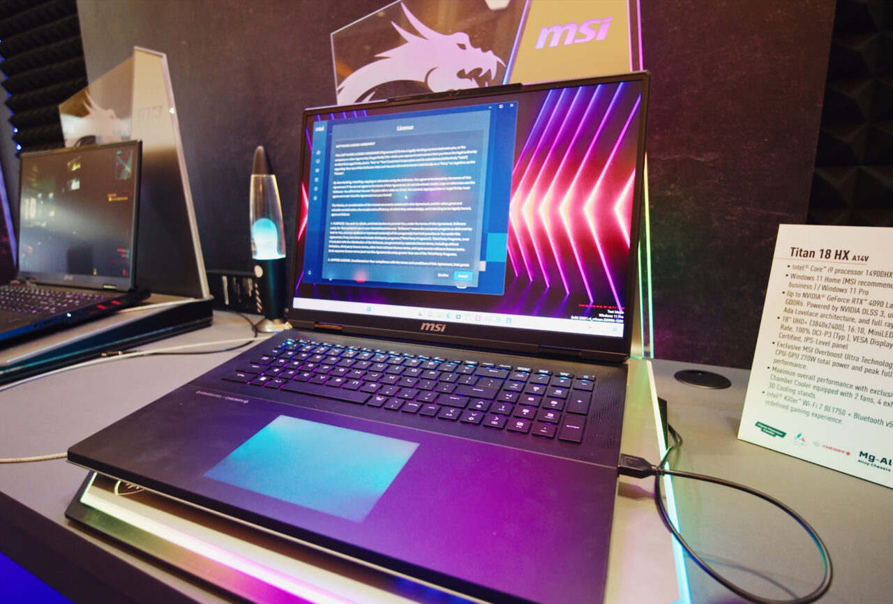 Laptop gamingowy MSI z podświetlaną klawiaturą na tle promocyjnego banera, obok opis modelu na stojaku informacyjnym.